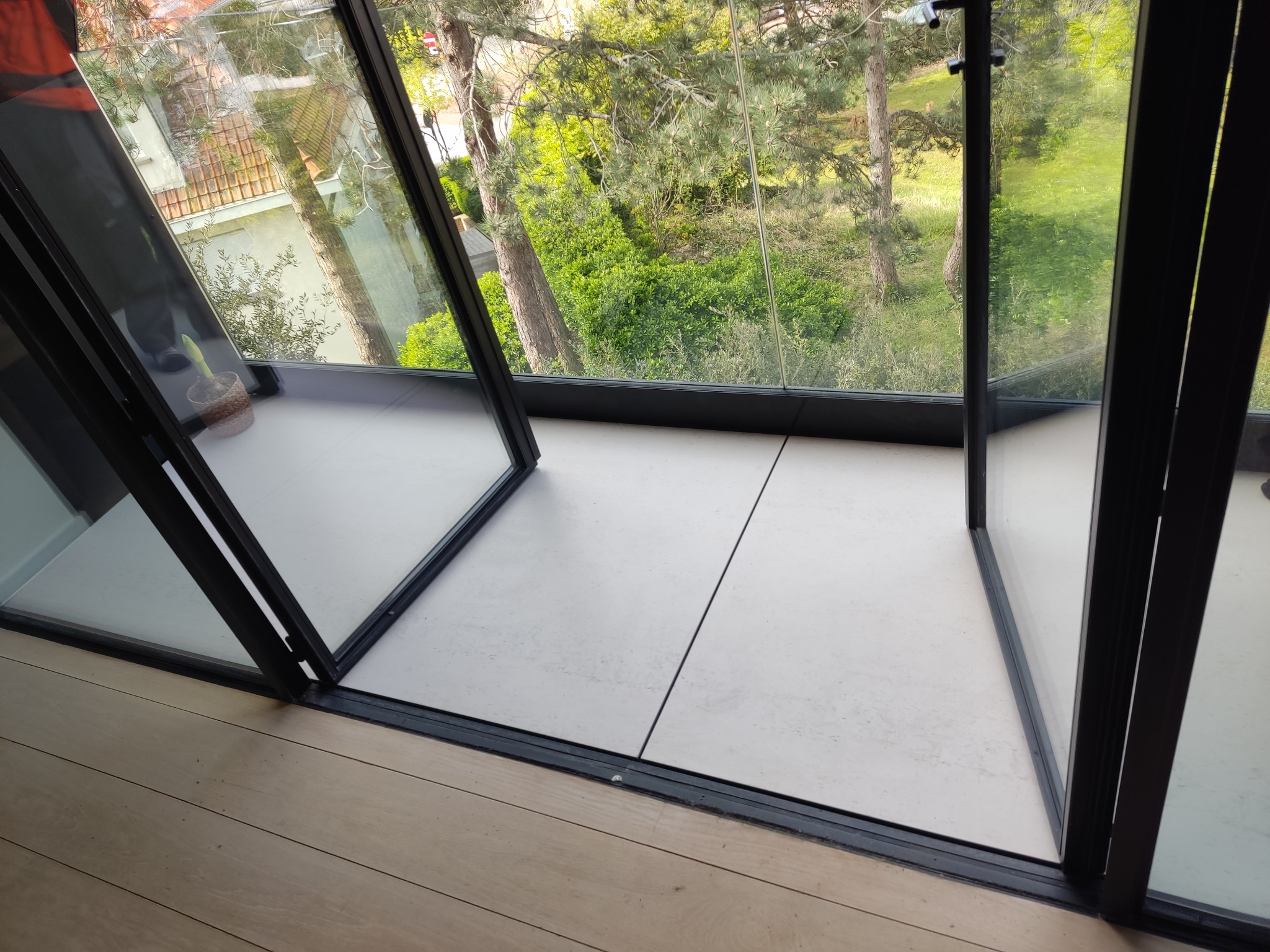 Nowoczesne, przesuwne okno z czarnymi ramami, otwierające się na zewnątrz, z widokiem na bujną zieleń. Wnętrze wykończone jasnymi, gładkimi płytami podłogowymi i drewnianą podłogą.