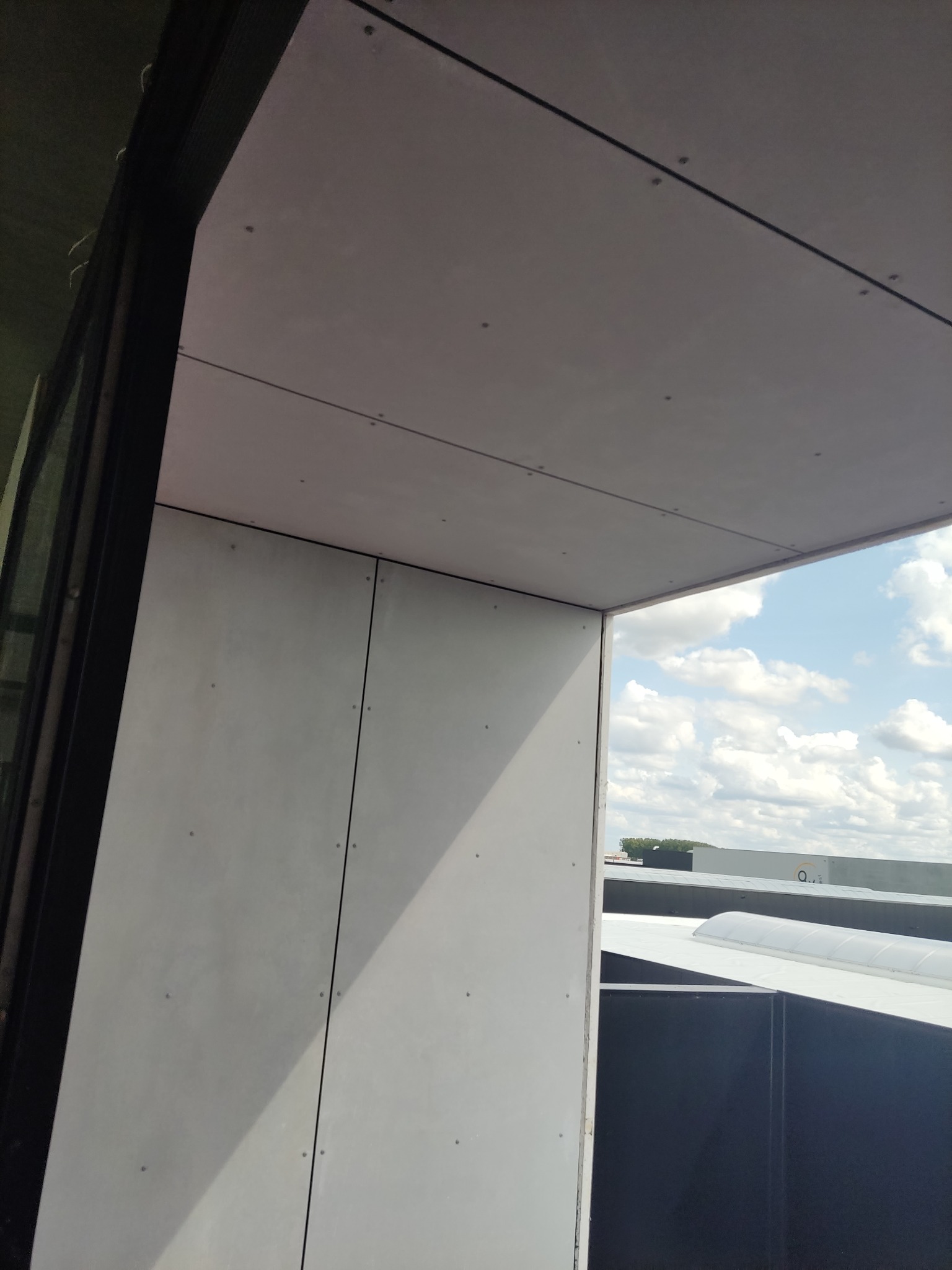 Wykończona ściana i sufit z płyt karton gips, widoczne łączenia i wkręty, fragment okna i widok na zabudowania przemysłowe w tle.