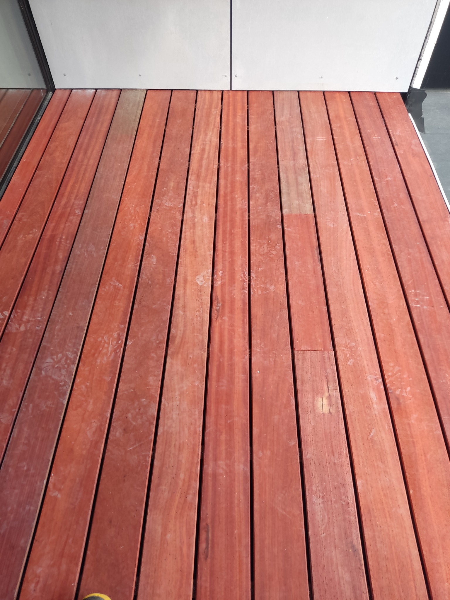 Ułożony parkiet z desek o ciepłej, czerwono-brązowej barwie, widoczne ślady użytkowania i delikatne zabrudzenia, przy ścianie z szarych płyt.