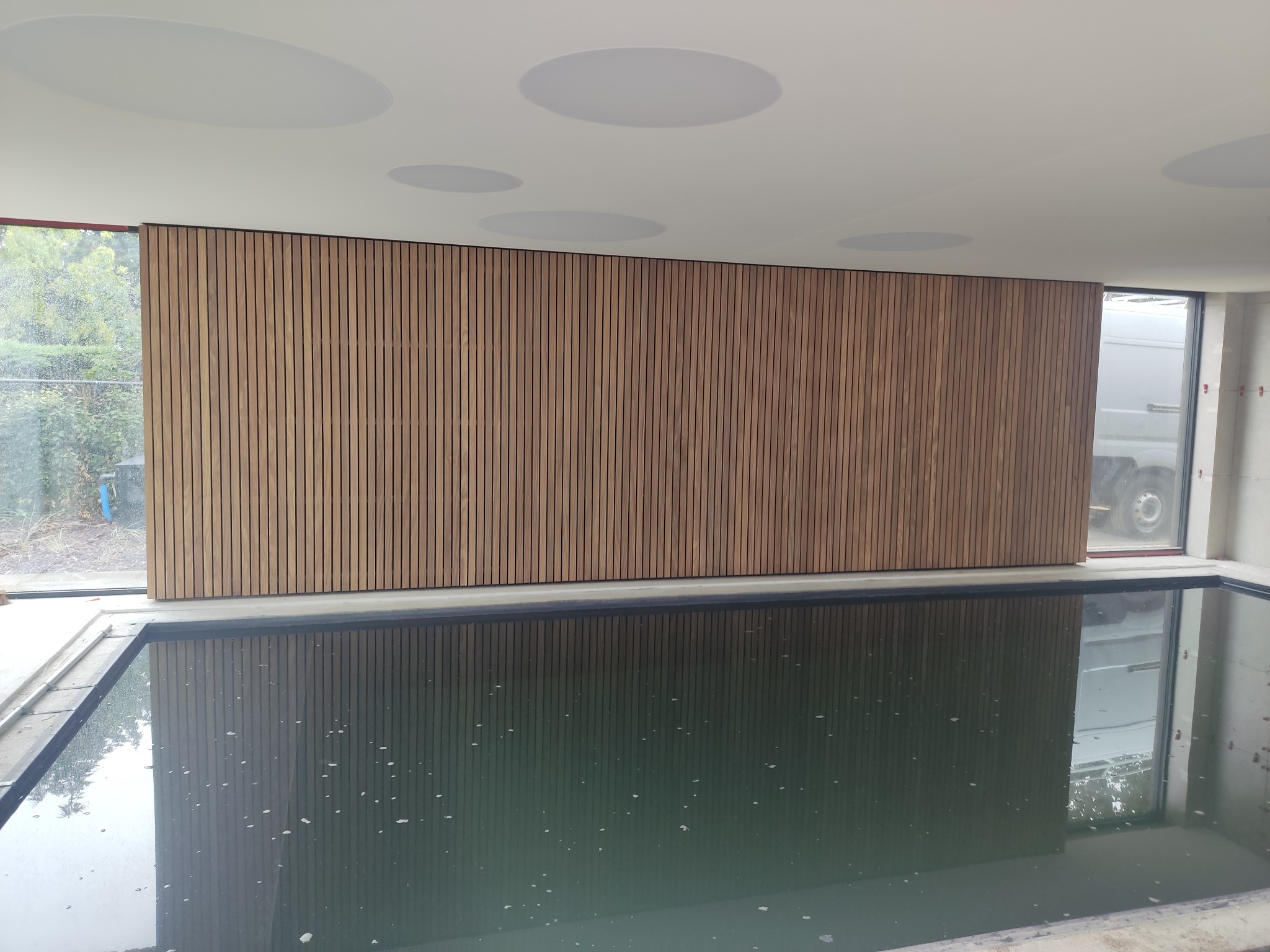 Drewniana okładzina ścienna z pionowych listew w pomieszczeniu z basenem, odbicie okładziny w wodzie, okrągłe głośniki sufitowe.