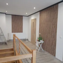 Korytarz z drewnianymi lamelami na ścianach, widok z góry schodów z drewnianą balustradą, białe ściany z czarnymi listwami przysufitowymi, drabina aluminiowa w tle.