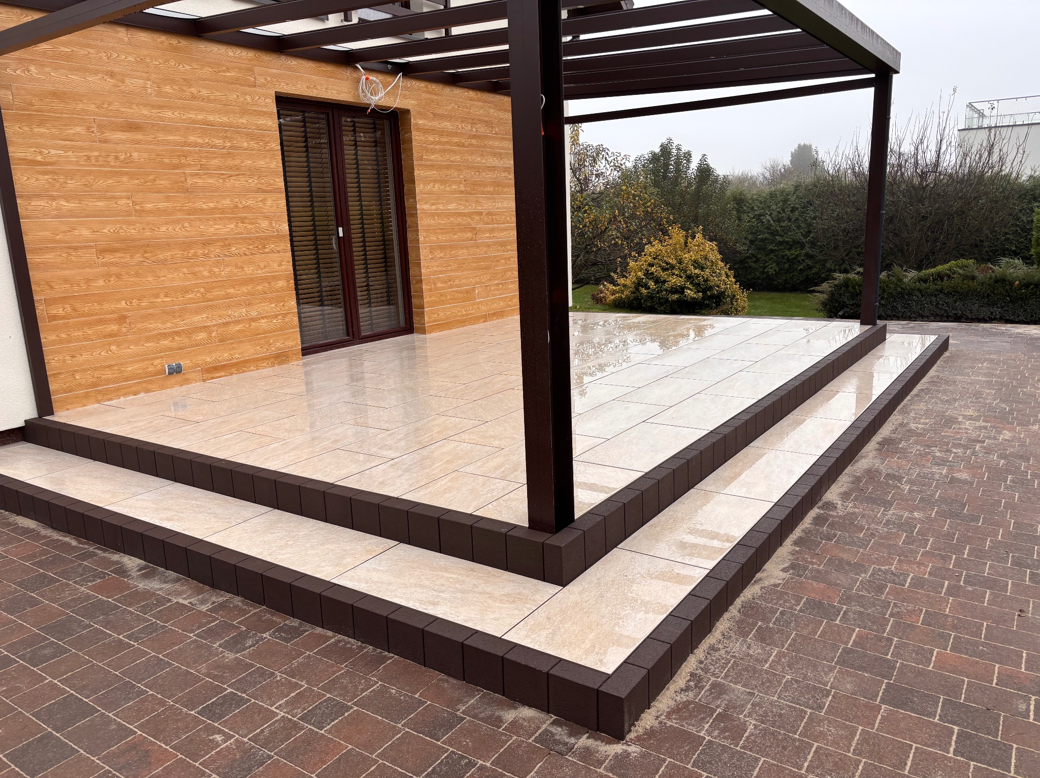 Wykończony taras z jasnymi płytkami imitującymi kamień, obramowany ciemnobrązową kostką, z widoczną pergolą i fragmentem elewacji budynku z drewnopodobną okładziną, oraz fragmentem nawierzchni...