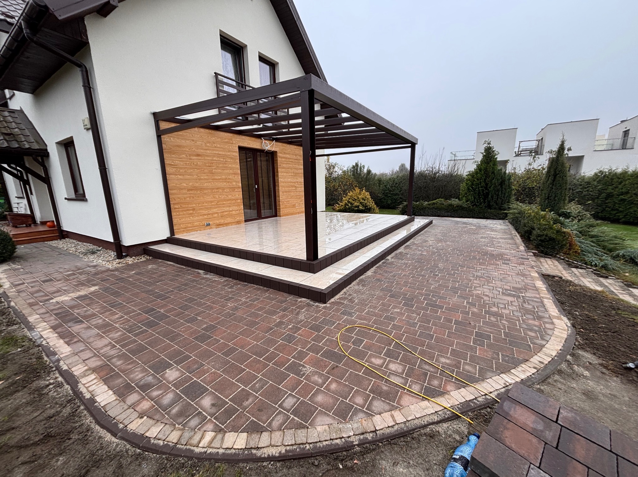Ułożona kostka brukowa w odcieniach czerwieni i brązu, z ciemniejszym obrzeżem, prowadząca do tarasu z pergolą przy nowoczesnym domu. Na ziemi leży żółty wąż ogrodowy i stos kostki brukowej.