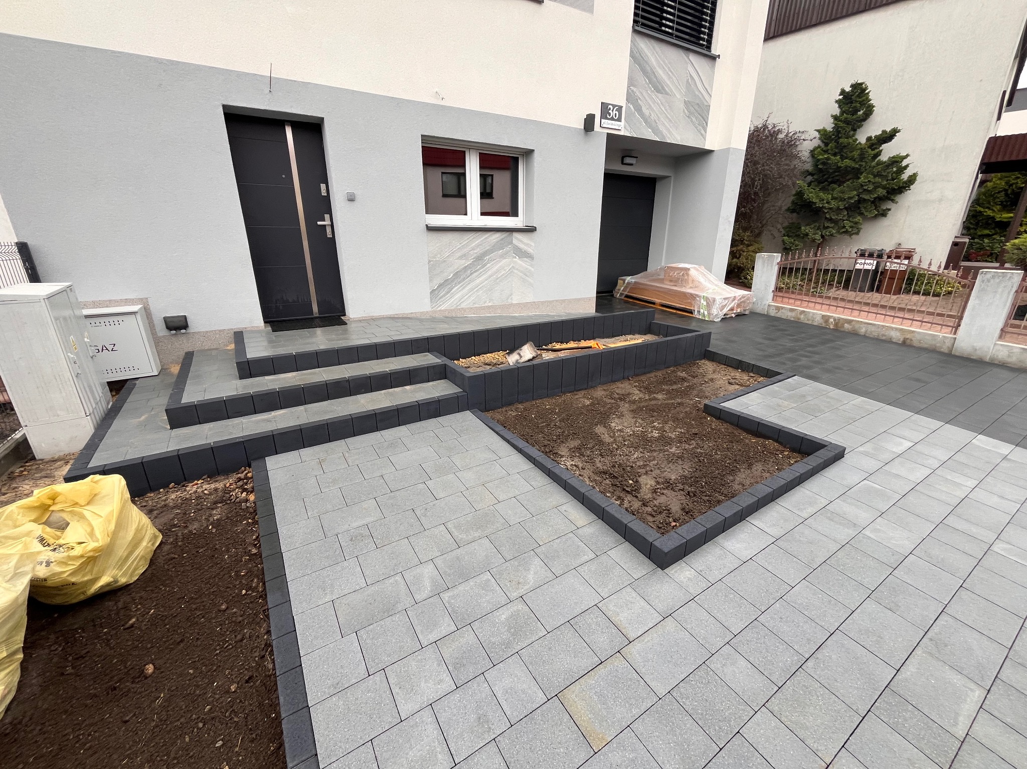 Nowoczesne wejście do domu z szarych kostek brukowych z ciemnoszarym obramowaniem, schody i rabaty w trakcie aranżacji, widoczne worki z materiałami budowlanymi.