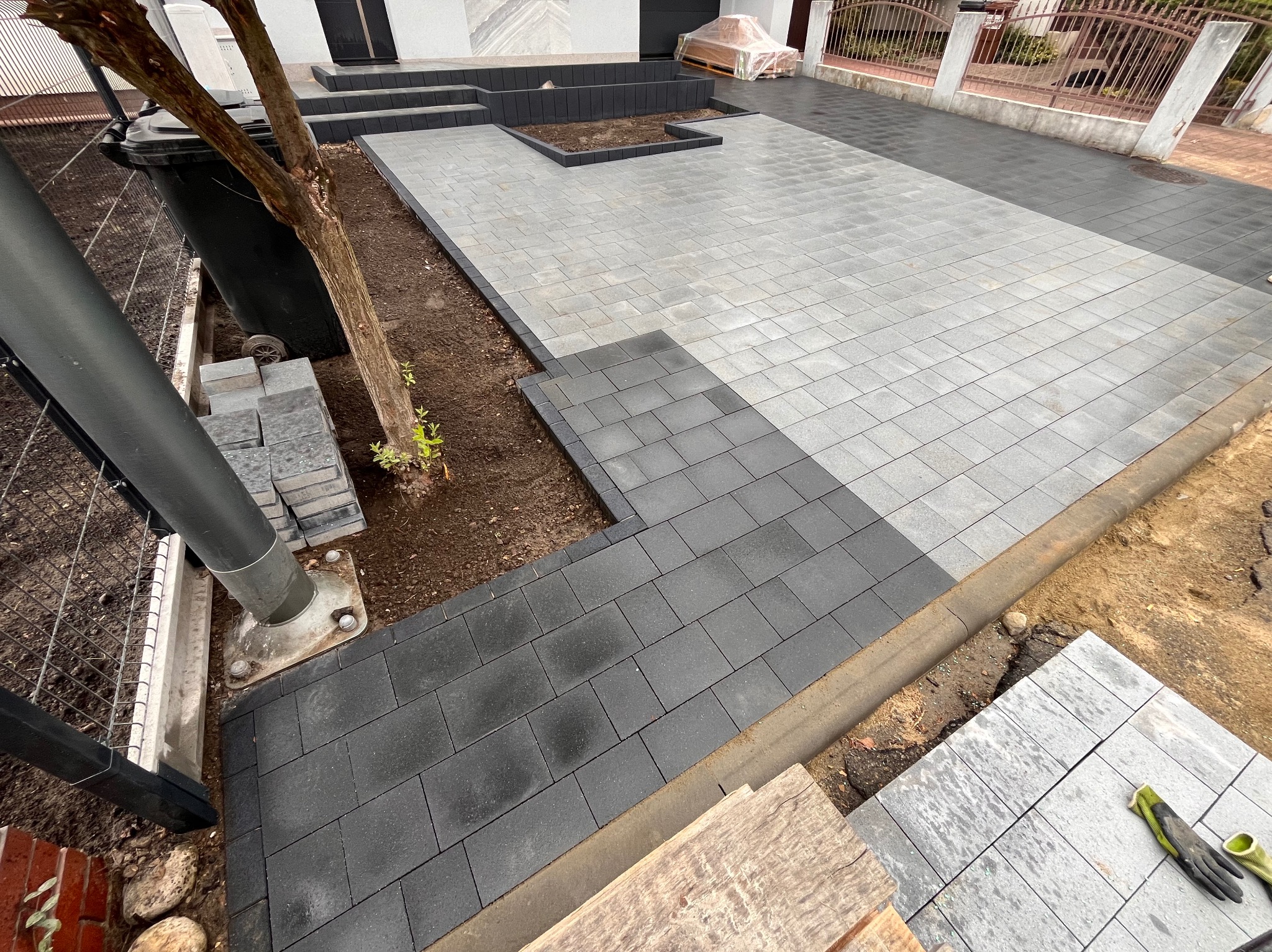 Ułożona szara kostka brukowa przed domem z czarnymi obrzeżami, widoczny fragment ogrodzenia i drzewo, po lewej stronie czarny kontener na śmieci.