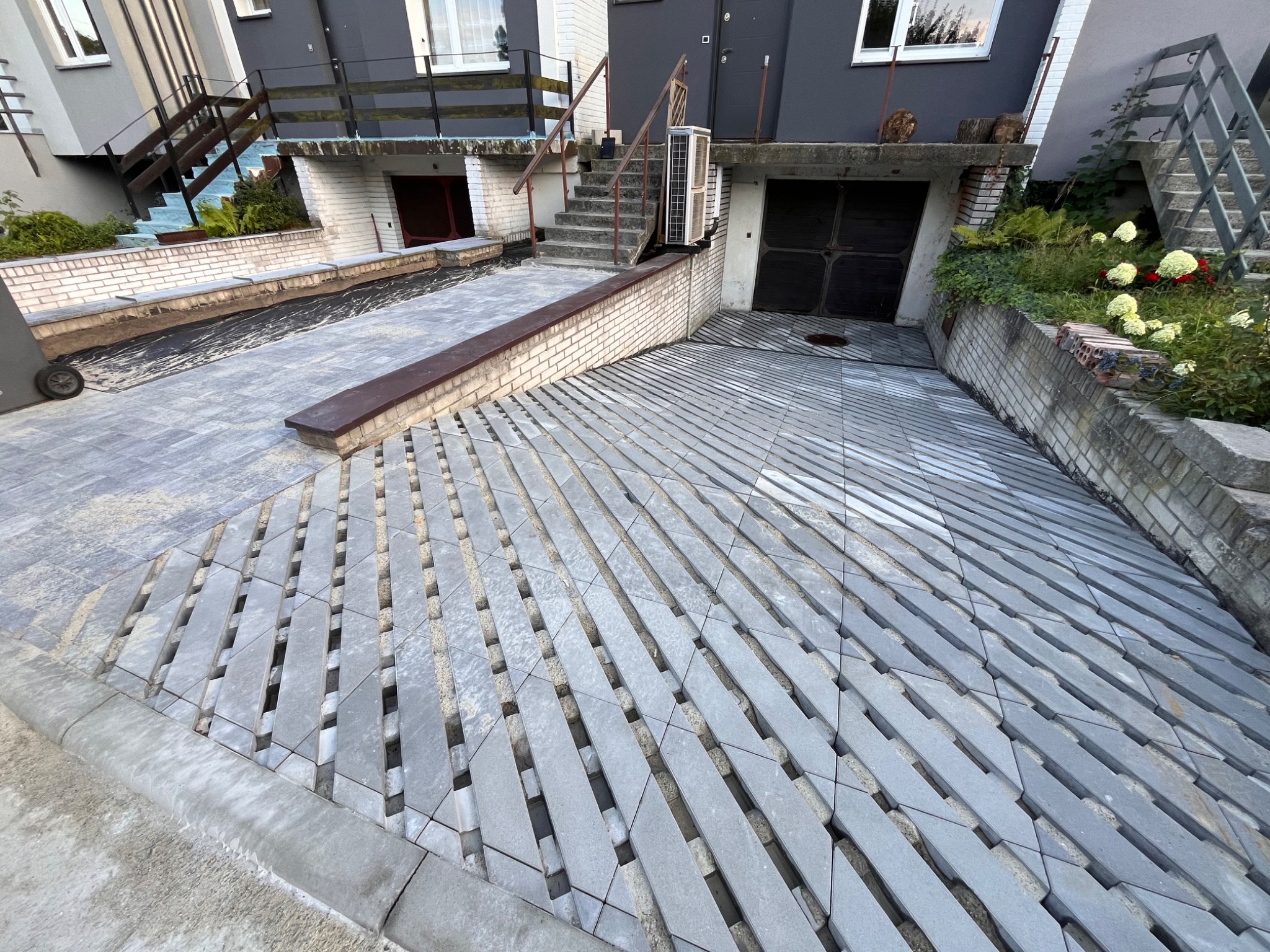 Ukośny wzór szarych płyt chodnikowych z widocznymi szczelinami wypełnionymi piaskiem, prowadzący do garażu w nowoczesnym domu z szarą elewacją i ceglanymi elementami.