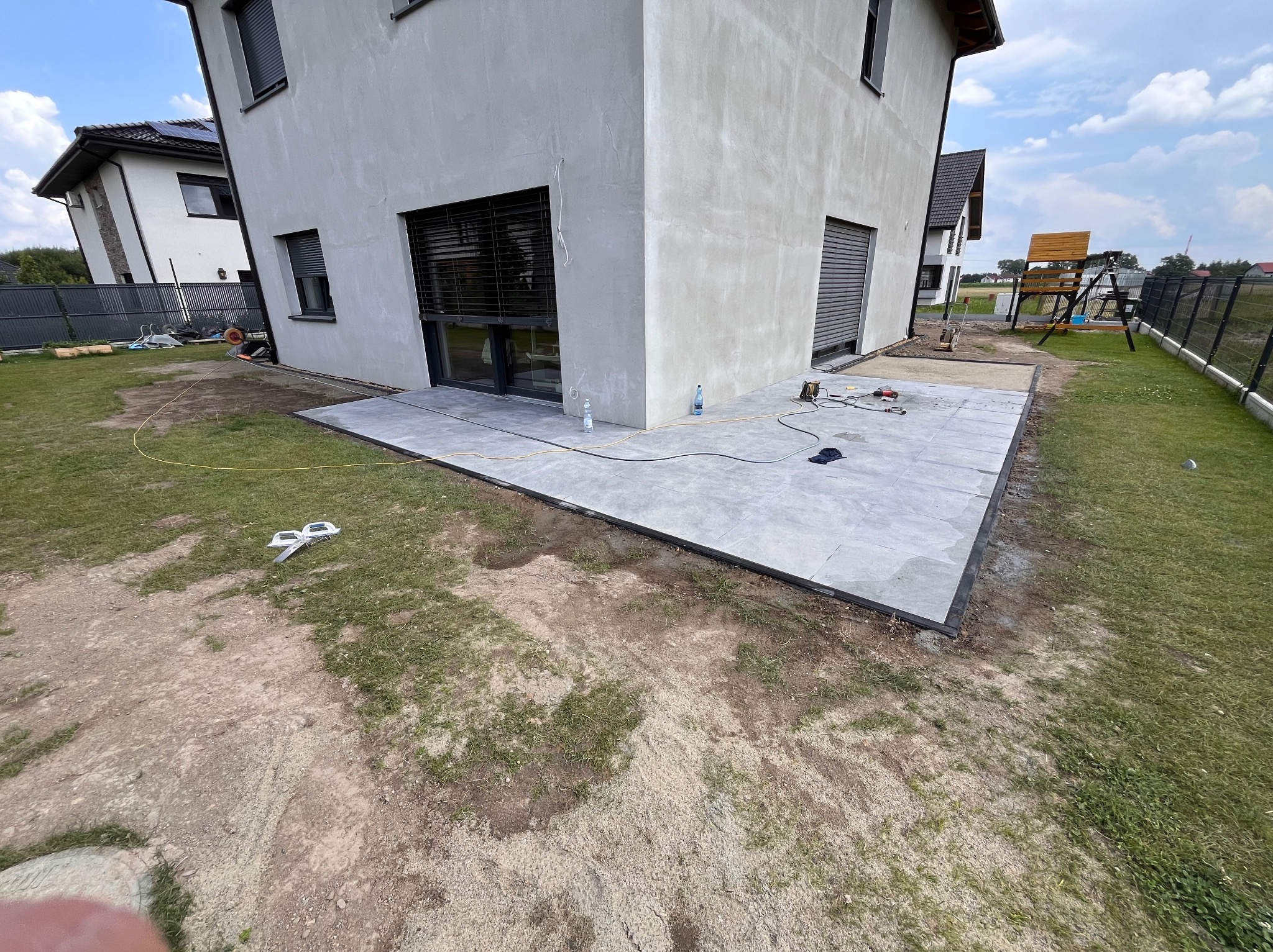 Nowoczesny taras z szarych płyt, obrzeża z czarnego tworzywa, narzędzia i butelki wody na powierzchni, dom z szarą elewacją w tle.