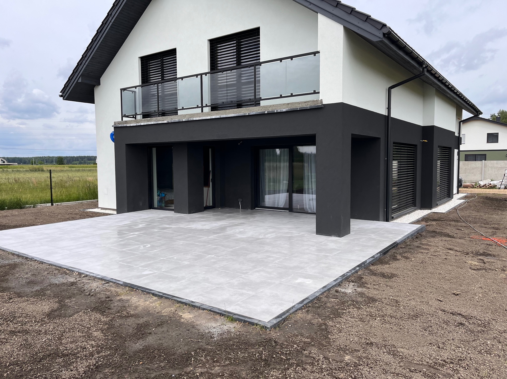 Nowoczesny taras z jasnoszarej kostki brukowej ułożonej przy dwukondygnacyjnym domu z balkonem i szklanymi balustradami. Widoczne obrzeża tarasu i przygotowany grunt wokół.