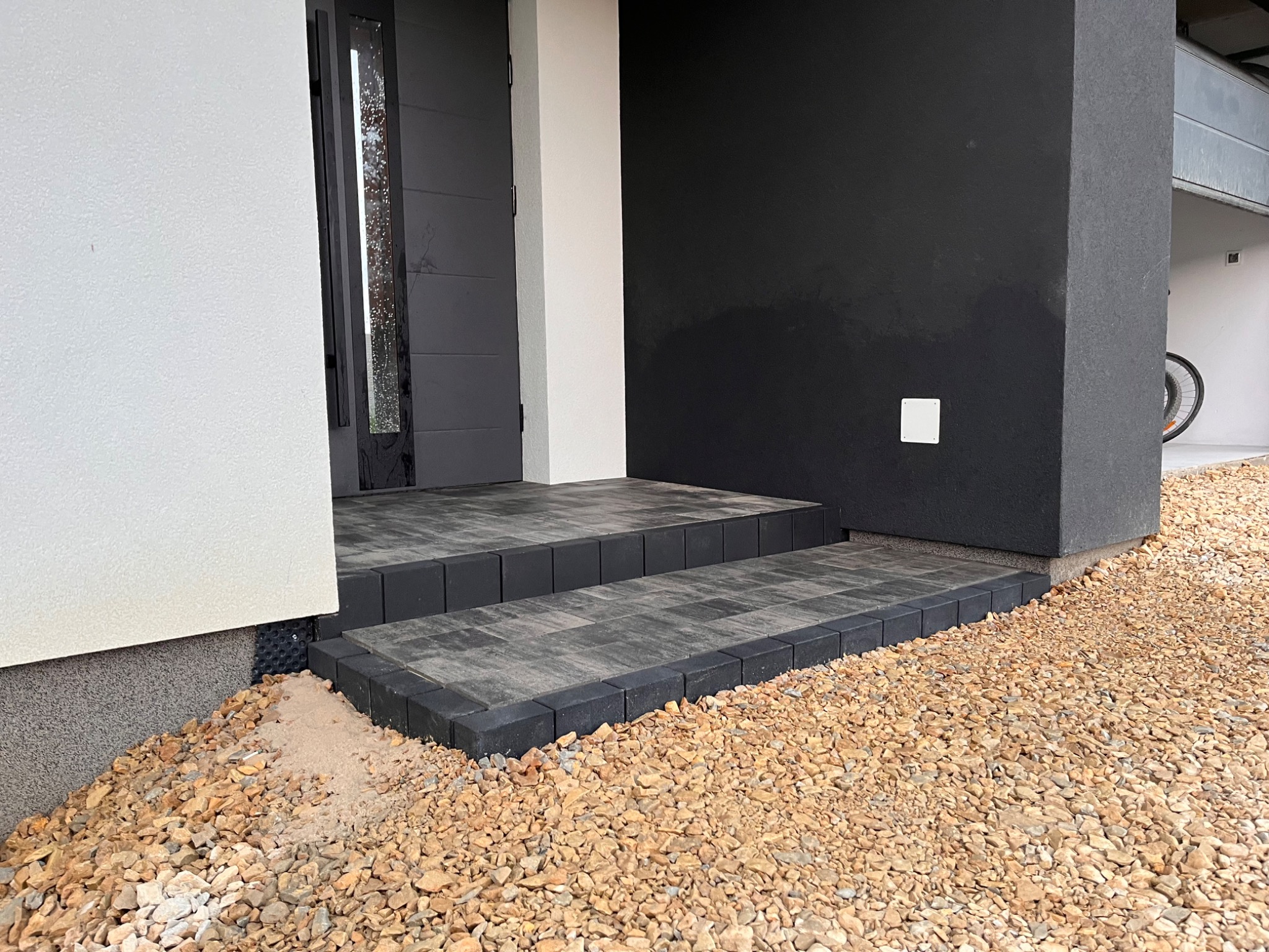 Schody wejściowe wyłożone szarą kostką brukową z czarnym obramowaniem, otoczone drobnym, brązowym żwirem, na tle nowoczesnego domu z czarno-białą elewacją.