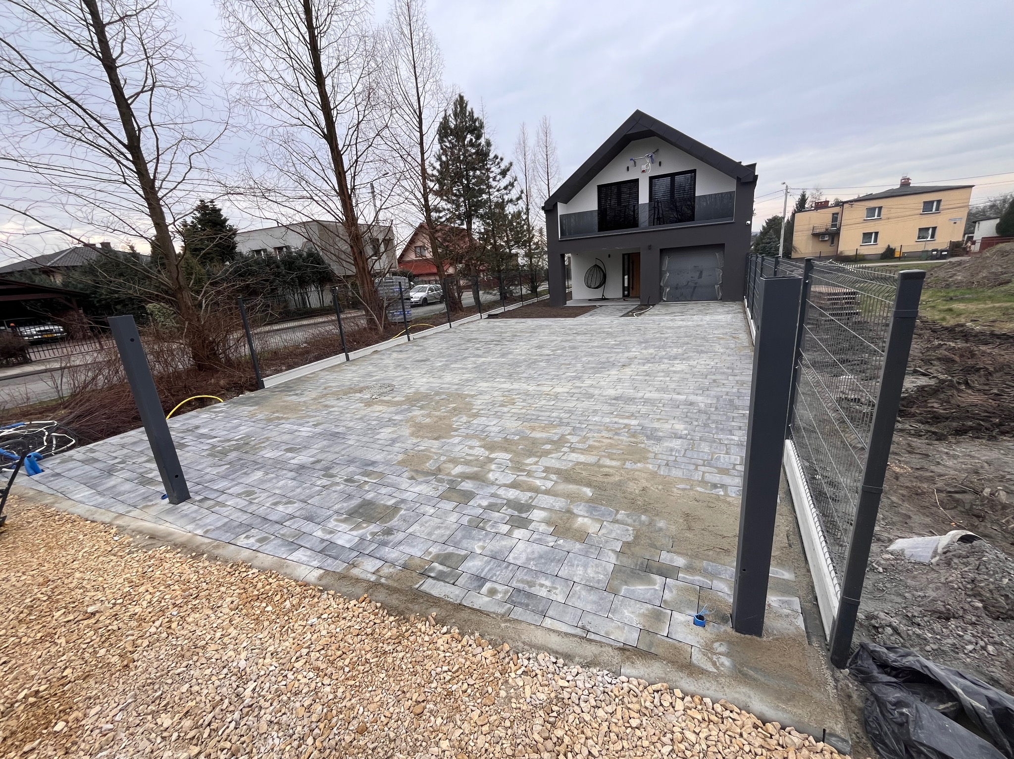 Ułożona szara kostka brukowa na podjeździe do nowoczesnego domu z szarą elewacją, widoczne słupki ogrodzeniowe i podsypka z kruszywa