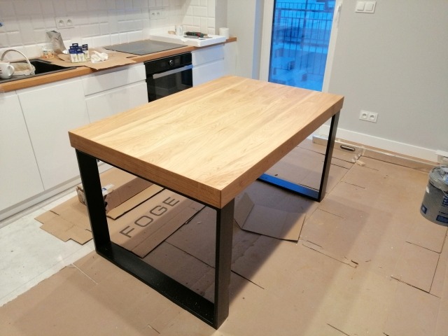 Nowoczesny stół z blatem z jasnego drewna i czarnymi, metalowymi nogami, stojący w kuchni w trakcie remontu, z widocznymi kartonami zabezpieczającymi podłogę.