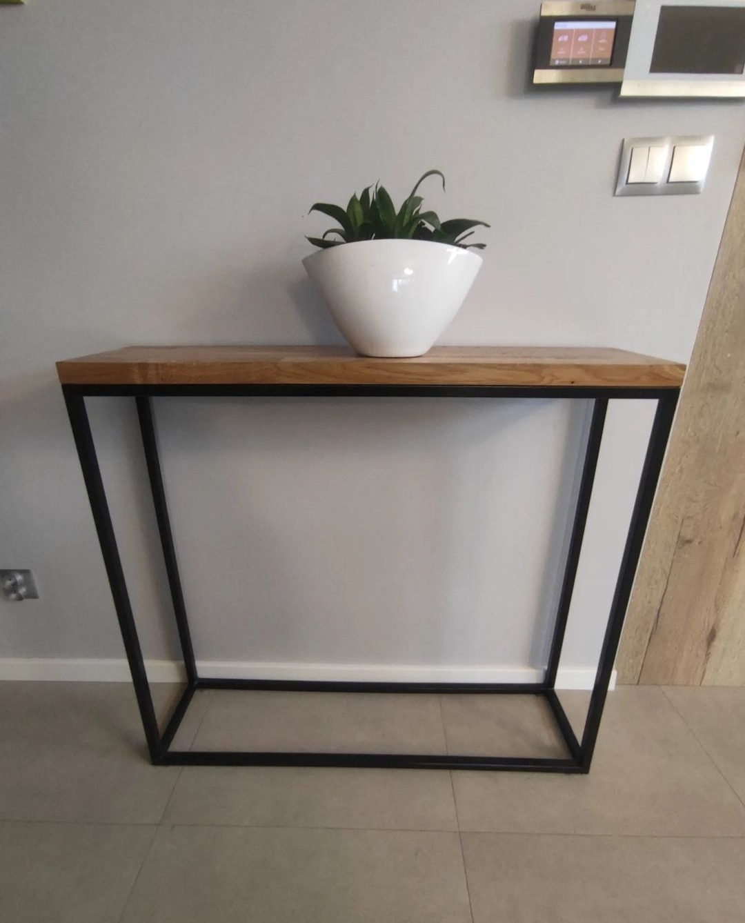 Nowoczesny stolik konsola z drewnianym blatem i czarną metalową ramą, na którym stoi biała donica z zieloną rośliną, na tle szarej ściany z panelem sterowania klimatyzacją i włącznikami światła.