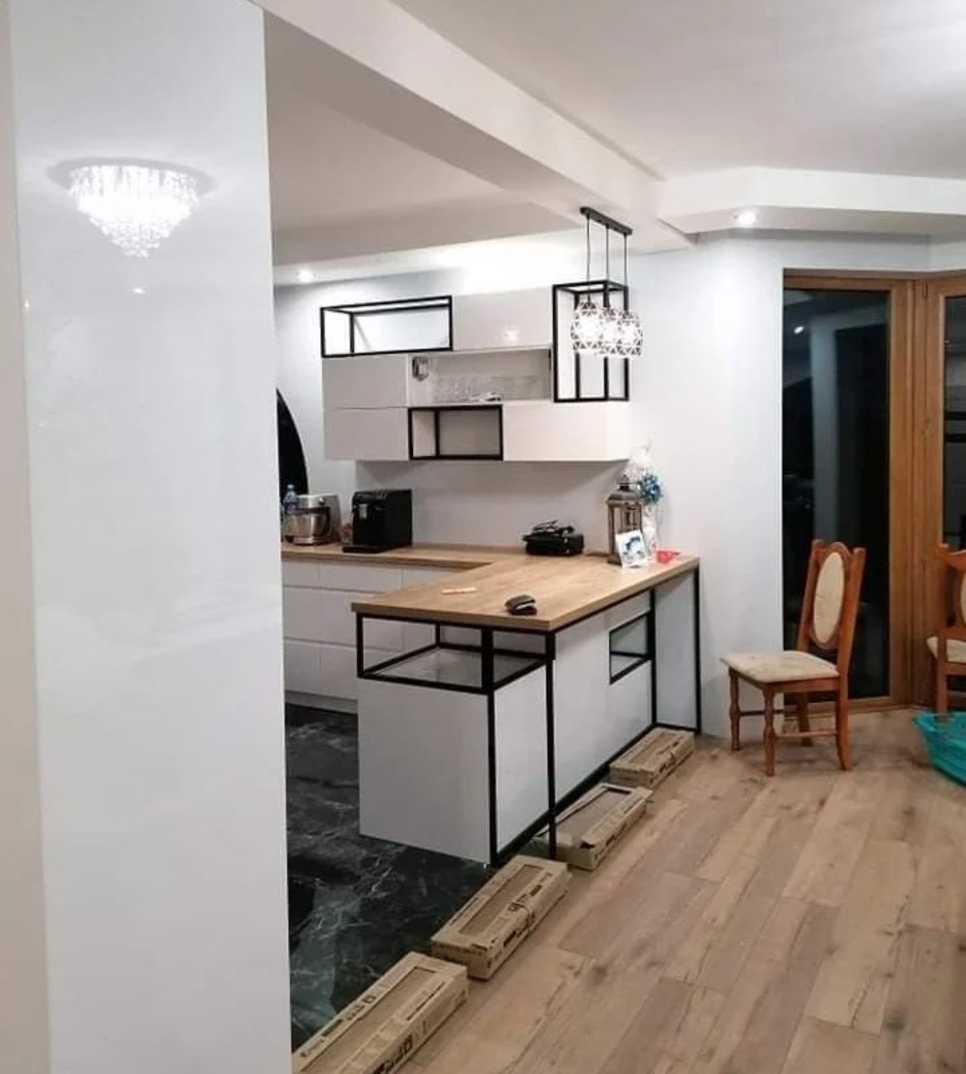 Nowoczesna kuchnia z wyspą o drewnianym blacie i czarnych metalowych elementach, widoczna także biała zabudowa kuchenna z otwartymi półkami i designerska lampa wisząca, podłoga wyłożona panelami...