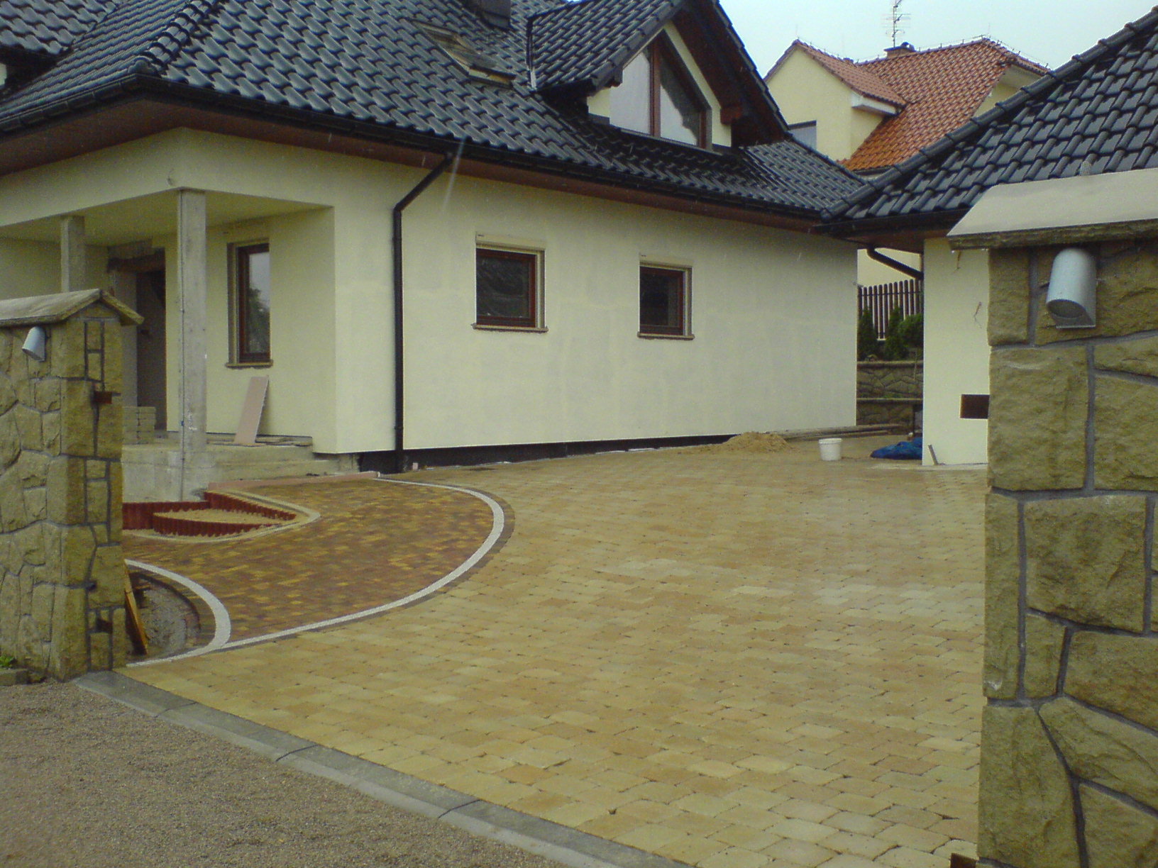 Żółta kostka brukowa ułożona na podjeździe przed domem, widok zza kamiennego słupka, w tle budynek w trakcie wykończenia elewacji.