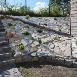 Skarpa umocniona jasnymi kamieniami z nasadzeniami niskich roślin ozdobnych, schody z kamienia prowadzą w górę, w tle siatka ogrodzeniowa i zieleń.