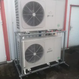 Dwie jednostki pompy ciepła Galmet Prima R32 zamontowane jedna nad drugą na metalowej konstrukcji wsporczej, na zewnątrz budynku o białej fasadzie z czerwonymi akcentami.