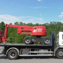 Czerwony podnośnik koszowy na lawecie ciężarówki pomocy drogowej, zabezpieczony łańcuchami, na tle zieleni.