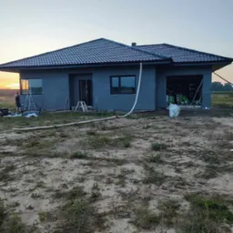 Dom jednorodzinny w trakcie ocieplania elewacji, widoczne płyty izolacyjne na ścianach zewnętrznych, rura spustowa, teren wokół posesji częściowo zagospodarowany, osoba w tle, zachód słońca.