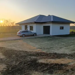 Nowoczesny dom jednorodzinny z szarą elewacją i grafitową dachówką w promieniach zachodzącego słońca, obok zaparkowany srebrny samochód kombi.