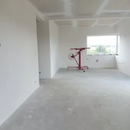 Wnętrze w trakcie prac wykończeniowych: sufit z widocznymi łączeniami płyt kartonowo-gipsowych przed szpachlowaniem, na podłodze czerwony podnośnik do płyt, ściany przygotowane do malowania, widok...