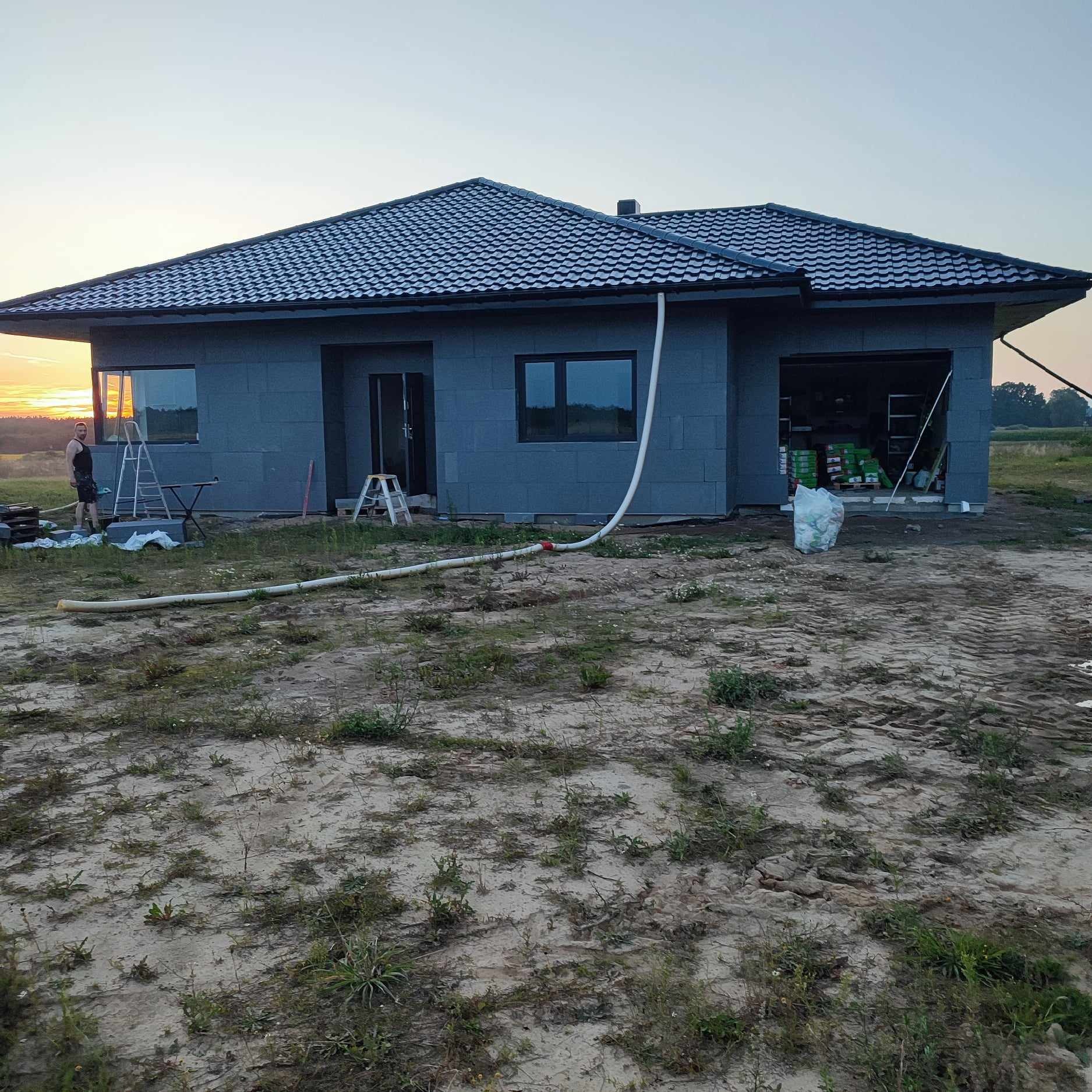 Dom jednorodzinny w trakcie ocieplania elewacji, widoczne płyty izolacyjne na ścianach zewnętrznych, rura spustowa, teren wokół posesji częściowo zagospodarowany, osoba w tle, zachód słońca.