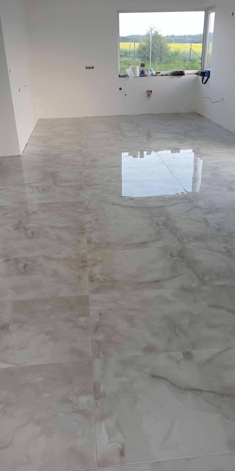 Błyszcząca, marmurkowa podłoga z płytek w nowym, jasnym pomieszczeniu z oknem, przez które widać pola.