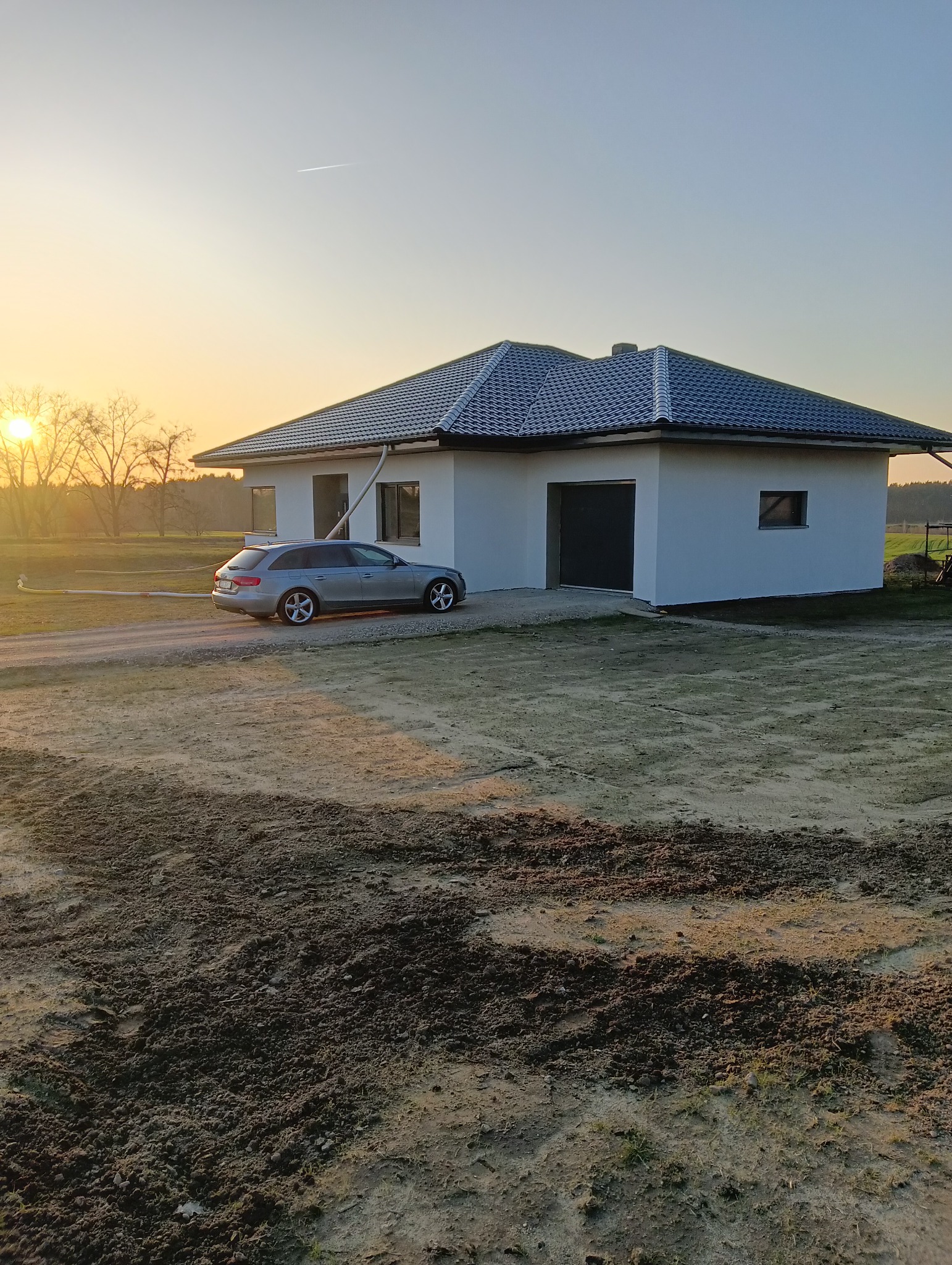 Nowoczesny dom jednorodzinny z szarą elewacją i grafitową dachówką w promieniach zachodzącego słońca, obok zaparkowany srebrny samochód kombi.