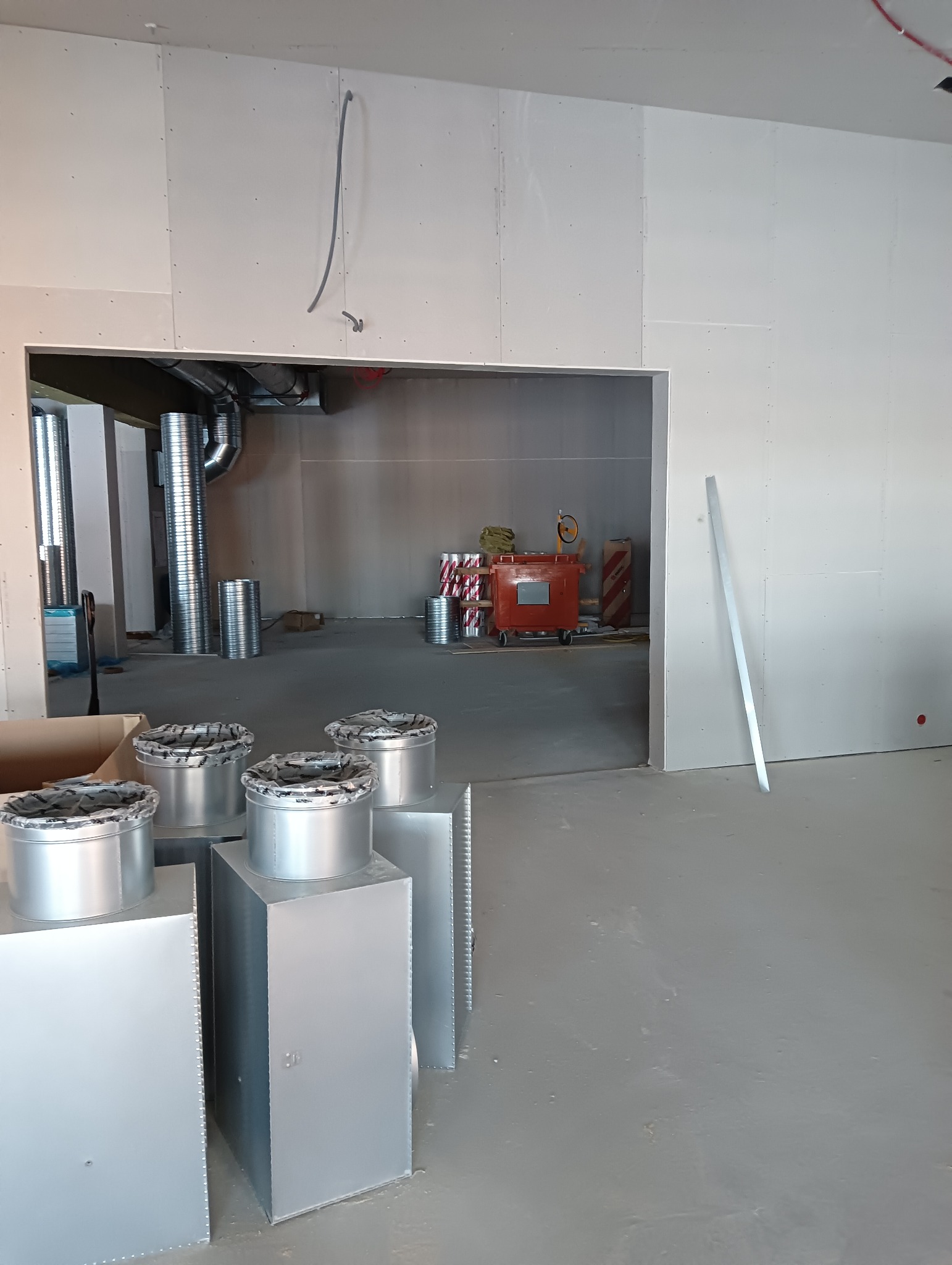 Wnętrze w trakcie prac wykończeniowych: widoczne płyty kartonowo-gipsowe na ścianach, elementy wentylacji w srebrnym kolorze oraz narzędzia i materiały budowlane w tle.