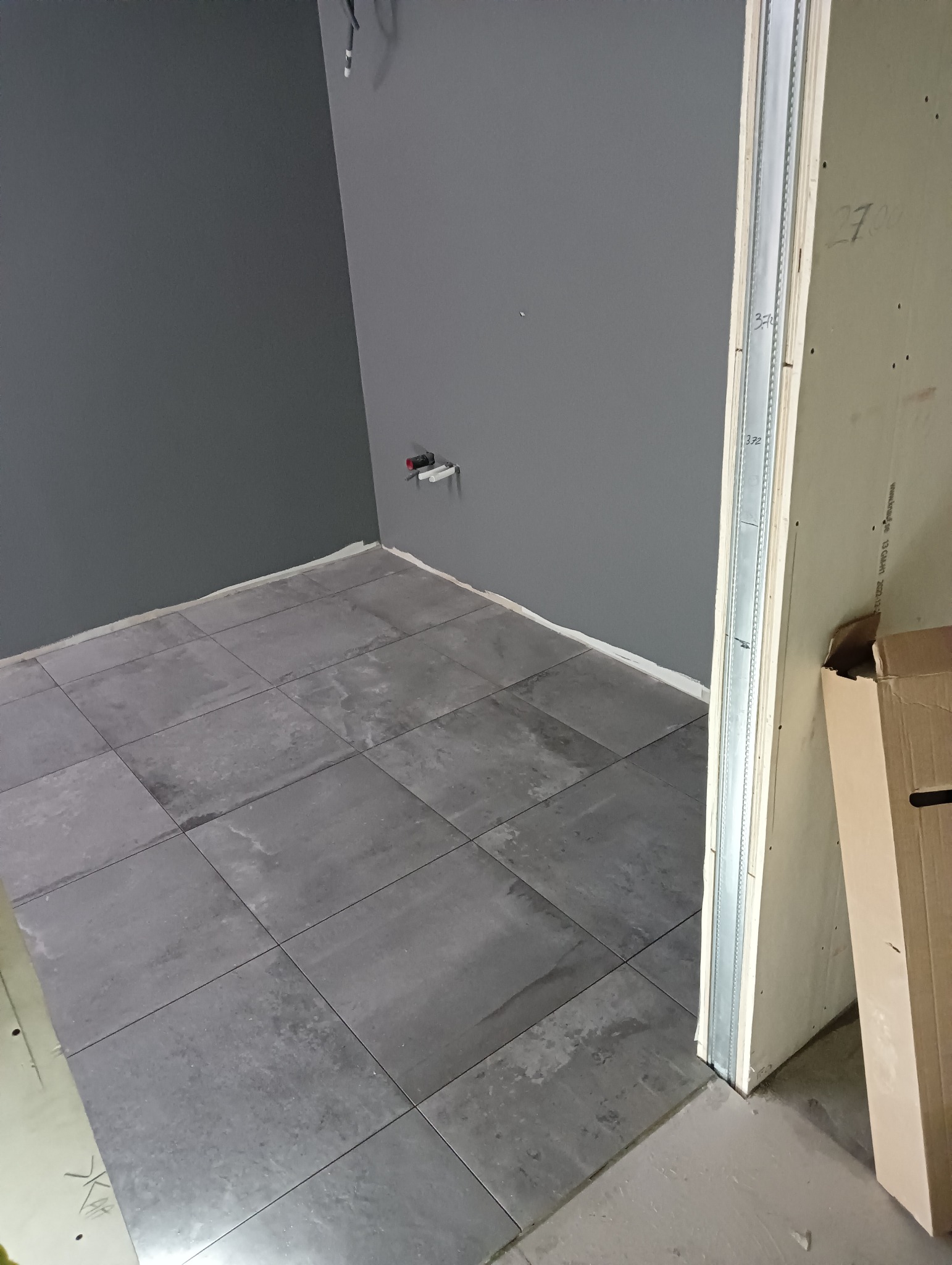 Szare płytki podłogowe w pomieszczeniu z szarą ścianą, widoczne fragmenty instalacji hydraulicznej i konstrukcji z płyty karton-gips w otworze drzwiowym.