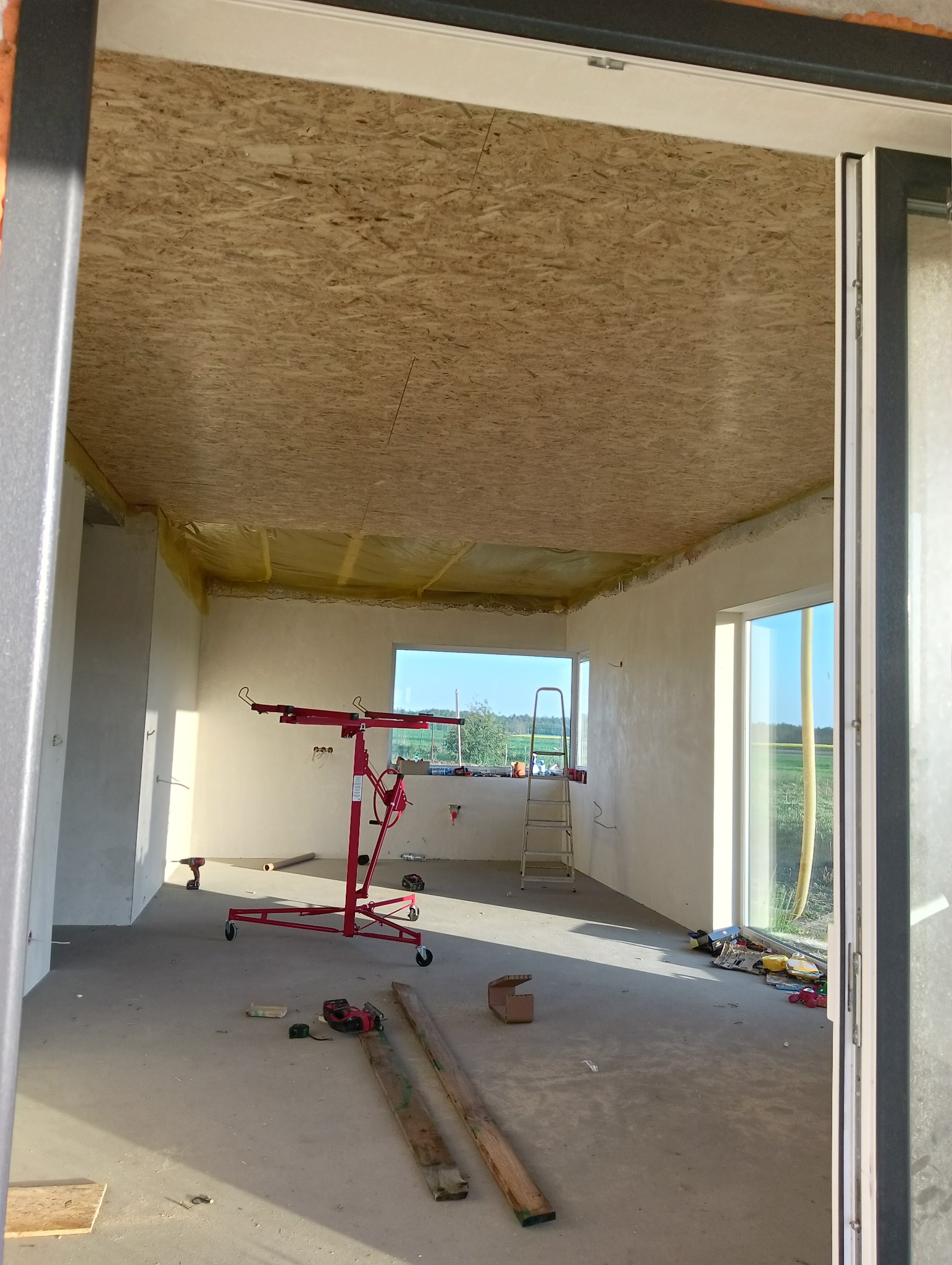 Wnętrze w trakcie prac wykończeniowych: sufit z płyt OSB, ściany przygotowane pod malowanie, widoczny podnośnik do płyt, drabina, narzędzia i materiały budowlane.