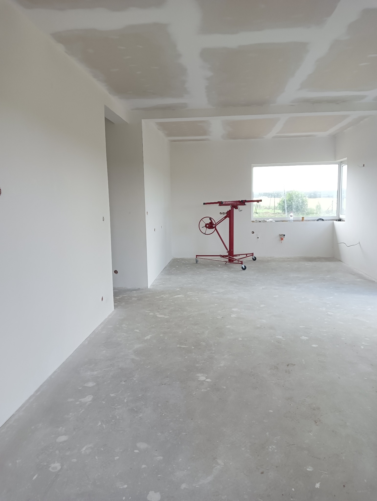 Wnętrze w trakcie prac wykończeniowych: sufit z widocznymi łączeniami płyt kartonowo-gipsowych przed szpachlowaniem, na podłodze czerwony podnośnik do płyt, ściany przygotowane do malowania, widok...