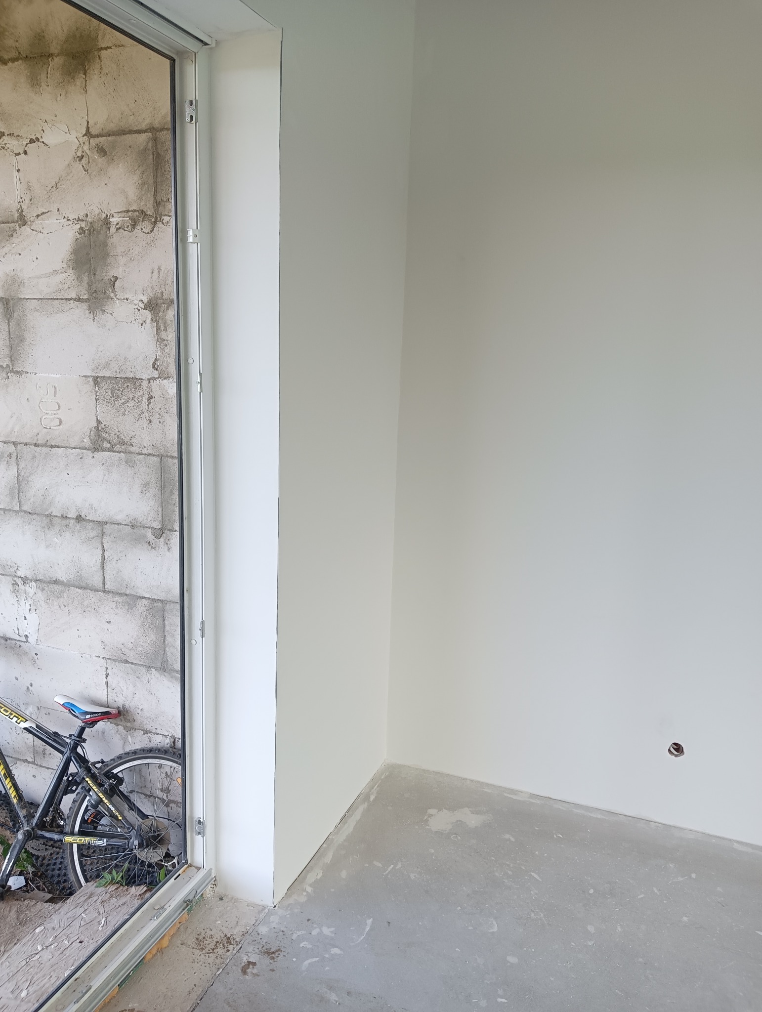 Wnętrze w trakcie remontu w Gryficach: pomalowane na biało ściany, surowa podłoga, widoczny fragment ściany z pustaków przez otwarte drzwi, oparty o ścianę rower.