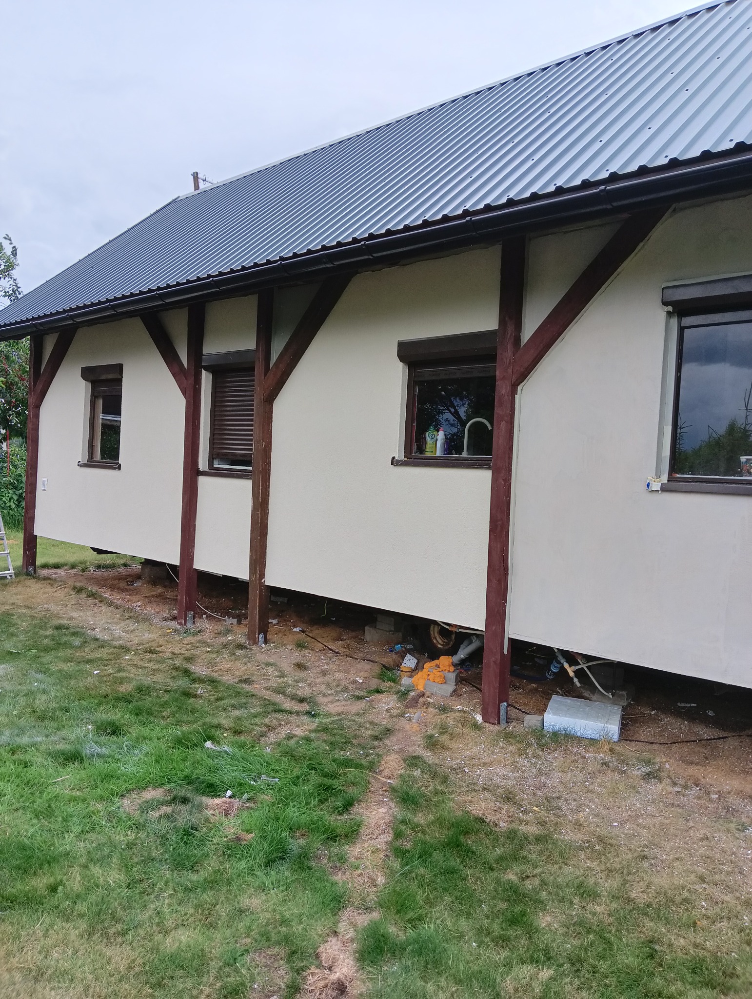 Wykończona elewacja domu z drewnianą konstrukcją wsporczą, widoczne okna z roletami i fragment trawnika.