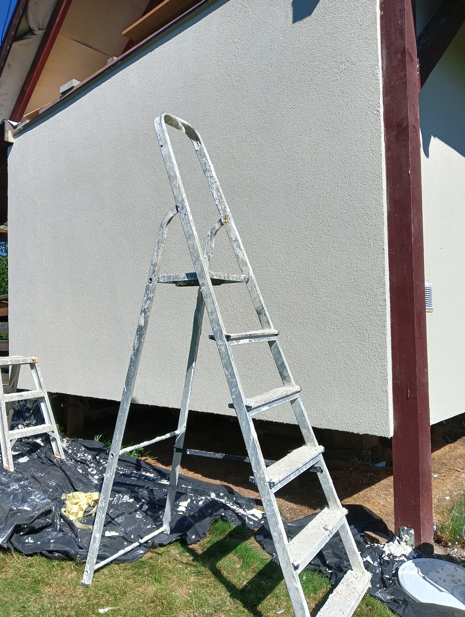 Świeżo otynkowana ściana budynku z widoczną drabiną aluminiową pokrytą resztkami tynku, zabezpieczona folią malarską na trawie.