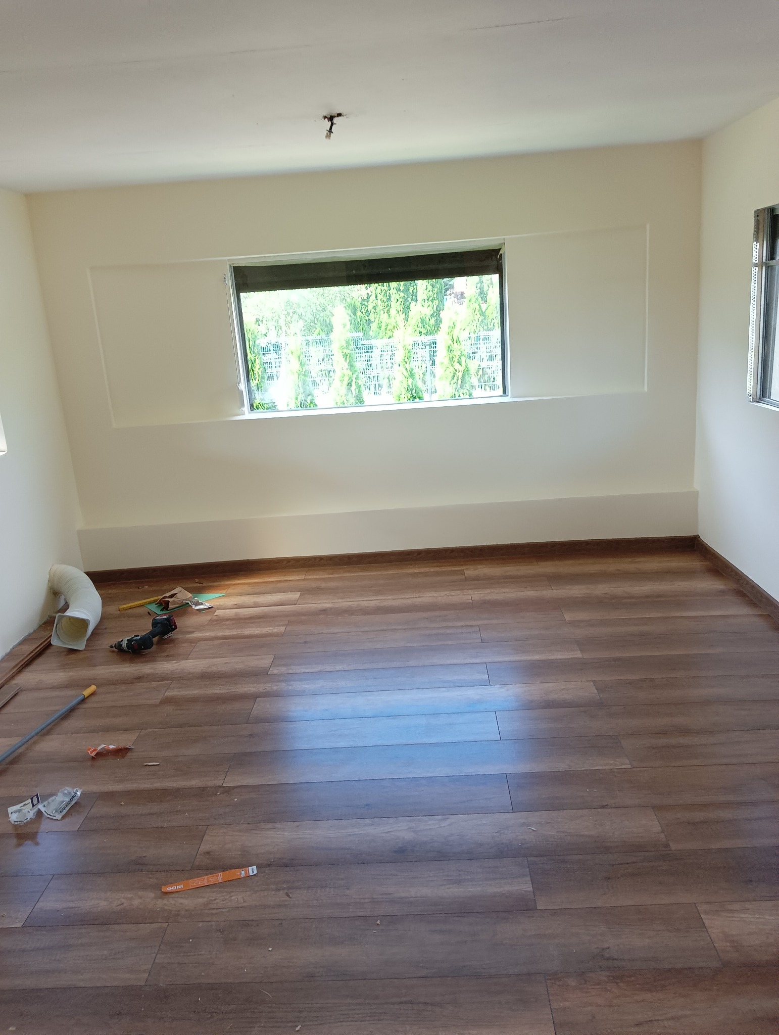Wnętrze pokoju po remoncie z drewnopodobną podłogą, świeżo pomalowanymi ścianami w odcieniu kremowym i widocznymi narzędziami budowlanymi na podłodze.