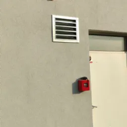 Szara ściana budynku z kratką wentylacyjną, czujnikiem pożarowym i rurą spustową, widoczne resztki styropianu po ociepleniu.