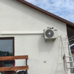 Zewnętrzna jednostka klimatyzacji marki AUX zamontowana na elewacji domu, z widoczną drabiną opartą o ścianę i fragmentem instalacji freonowej.