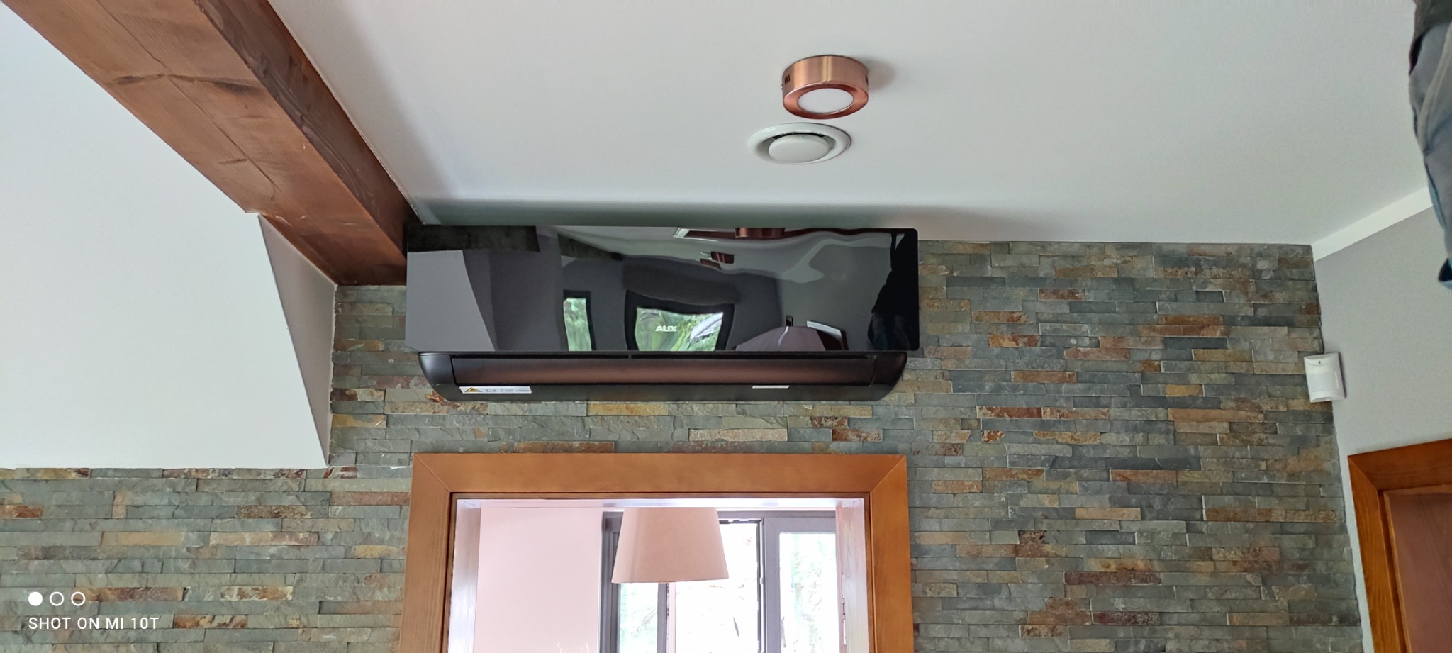 Czarny klimatyzator ścienny marki AUX zamontowany na ścianie z kamienia dekoracyjnego, widoczny fragment drewnianej belki stropowej i okrągły nawiewnik sufitowy