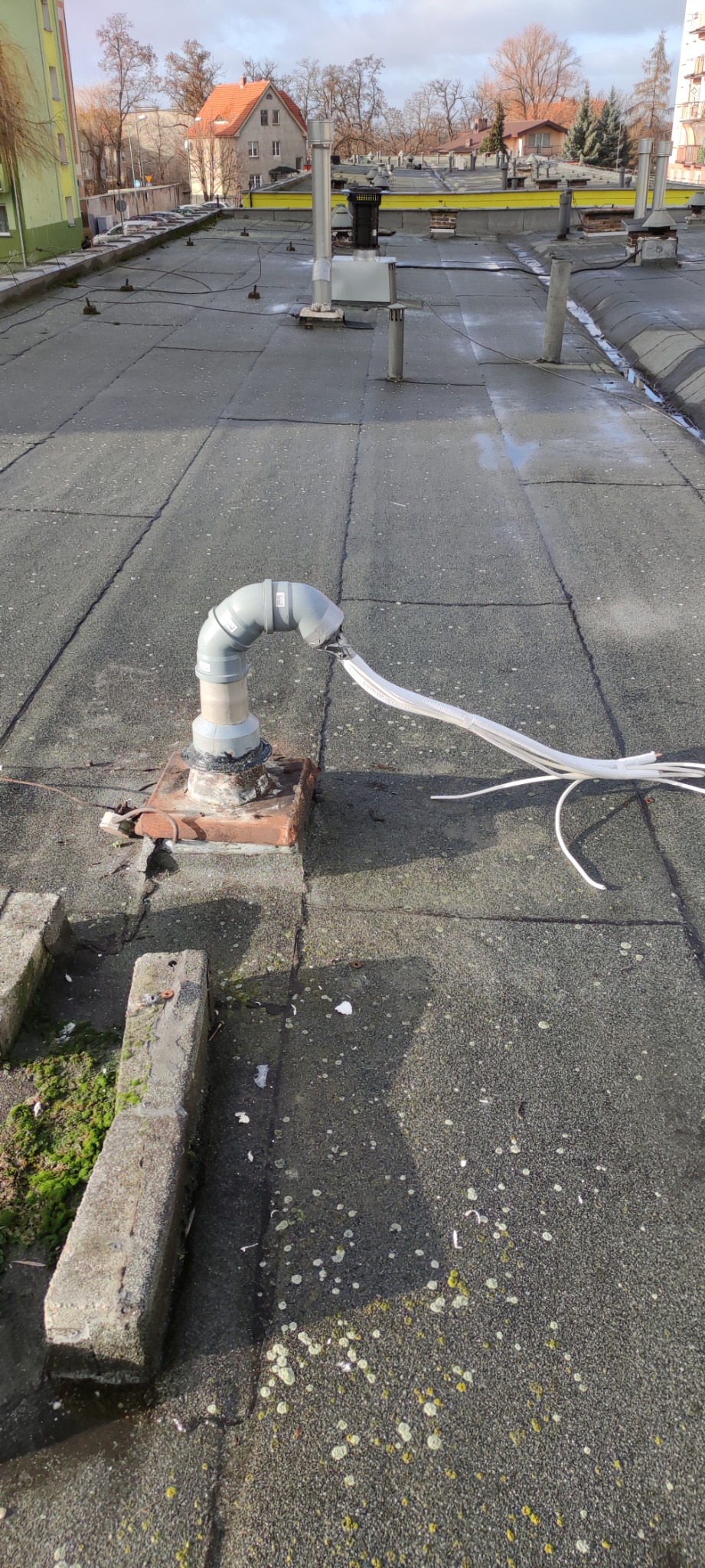 Szary przewód klimatyzacji wychodzący z rury na płaskim dachu pokrytym papą, widoczne budynki w tle.