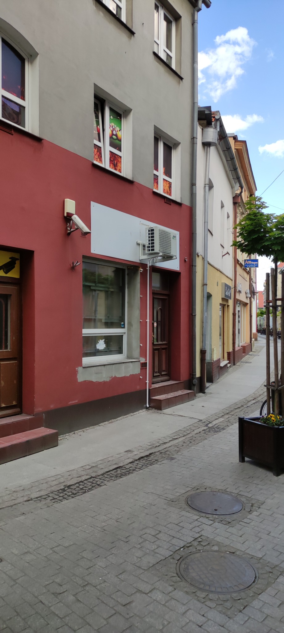 Zewnętrzna jednostka klimatyzacji zamontowana na czerwonej ścianie budynku przy wąskiej uliczce z brukowaną nawierzchnią.