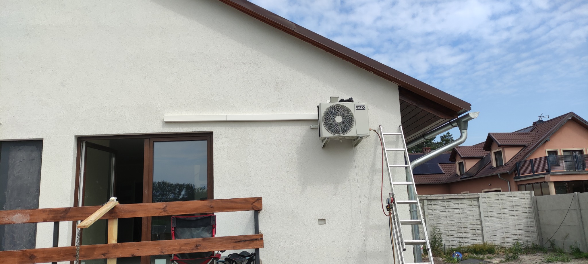 Zewnętrzna jednostka klimatyzacji marki AUX zamontowana na elewacji domu, z widoczną drabiną opartą o ścianę i fragmentem instalacji freonowej.