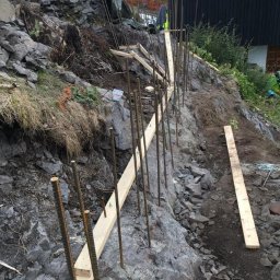 Solarispv - Przygotowanie zbrojenia i szalunku na skalistym zboczu pod budowę budynku żelbetowego, widoczne wystające pręty zbrojeniowe i drewniane elementy konstrukcyjne.