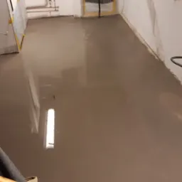 Świeżo wylana, mokra wylewka samopoziomująca na podłodze w pomieszczeniu z zabezpieczonymi folią drzwiami i widocznymi instalacjami na ścianie, z fragmentem narzędzia do rozprowadzania wylewki...