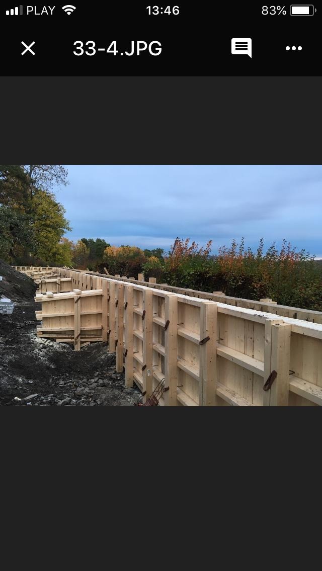 Szczegółowe ujęcie drewnianego szalunku fundamentowego na nierównym terenie, z widocznymi elementami mocującymi i jesienną roślinnością w tle pod pochmurnym niebem.
