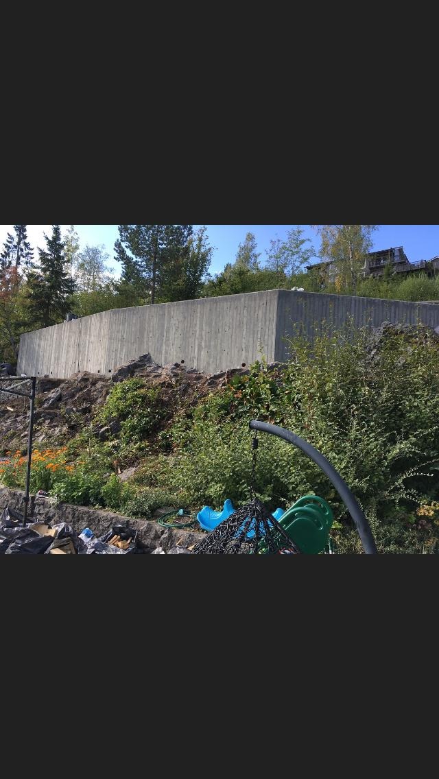 Surowa ściana oporowa z betonu na zboczu porośniętym roślinnością, z widocznymi elementami placu zabaw w dolnej części kadru.