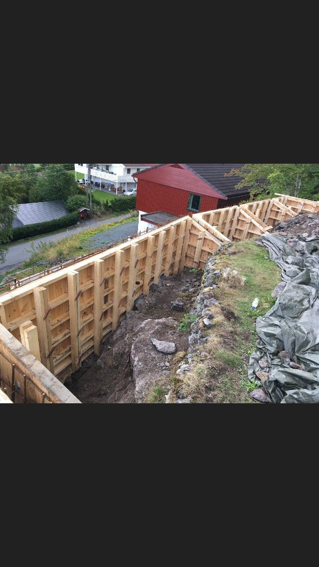 Szczegółowy widok szalunku z drewnianych desek, przygotowanego do wylania fundamentów na nierównym terenie, z widocznymi wystającymi prętami zbrojeniowymi i częściowo zakrytą folią skarpą.