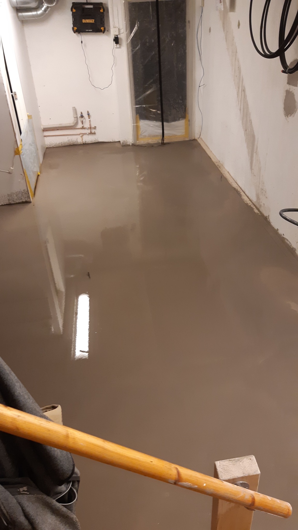 Świeżo wylana, mokra wylewka samopoziomująca na podłodze w pomieszczeniu z zabezpieczonymi folią drzwiami i widocznymi instalacjami na ścianie, z fragmentem narzędzia do rozprowadzania wylewki...