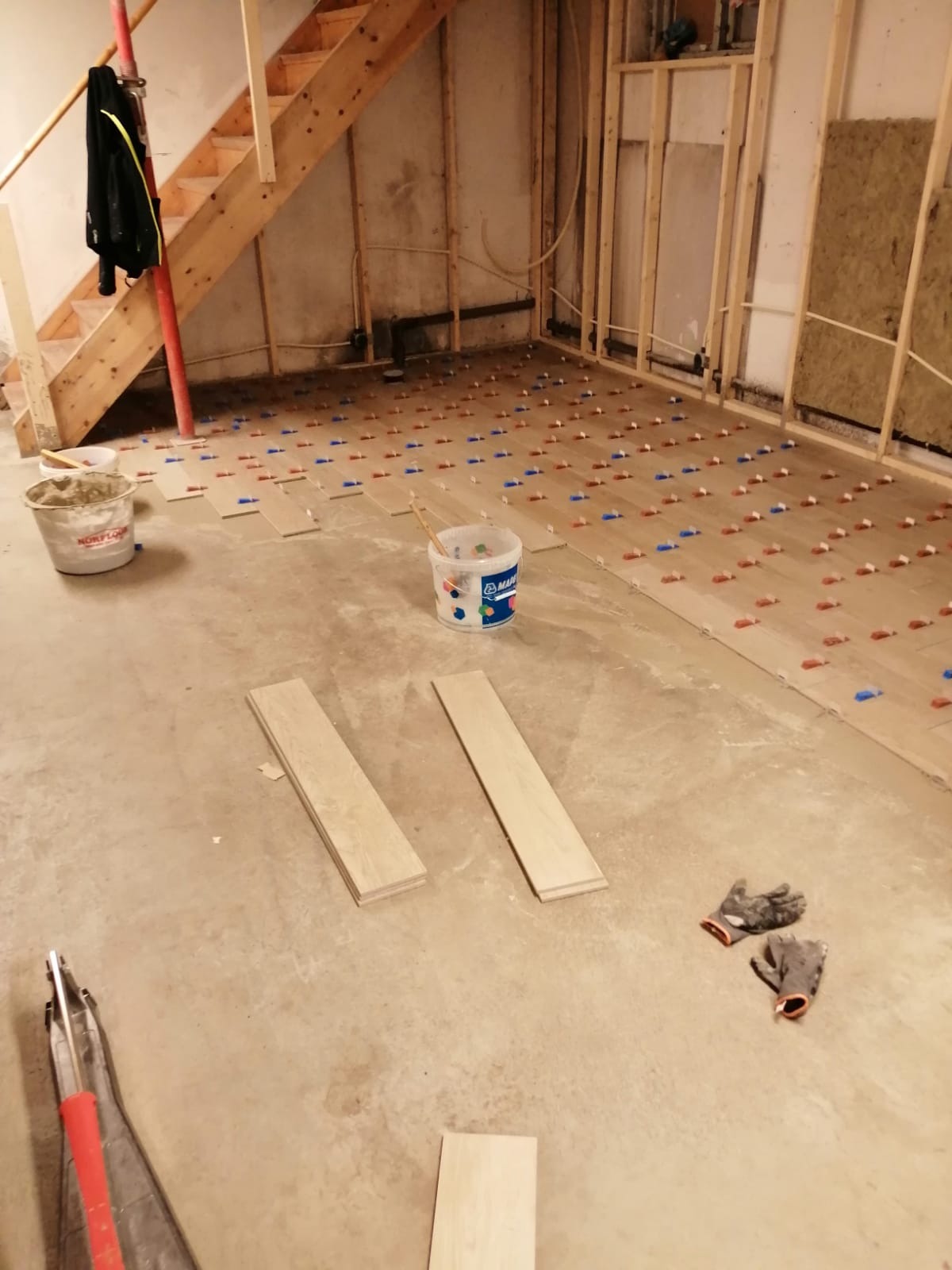 Układanie drewnianej podłogi: widoczne deski, wiadro z klejem Norfloor, wiadro z klejem Mapei, poziomica, rękawice robocze oraz krzyżyki dystansowe na podłodze w trakcie montażu.