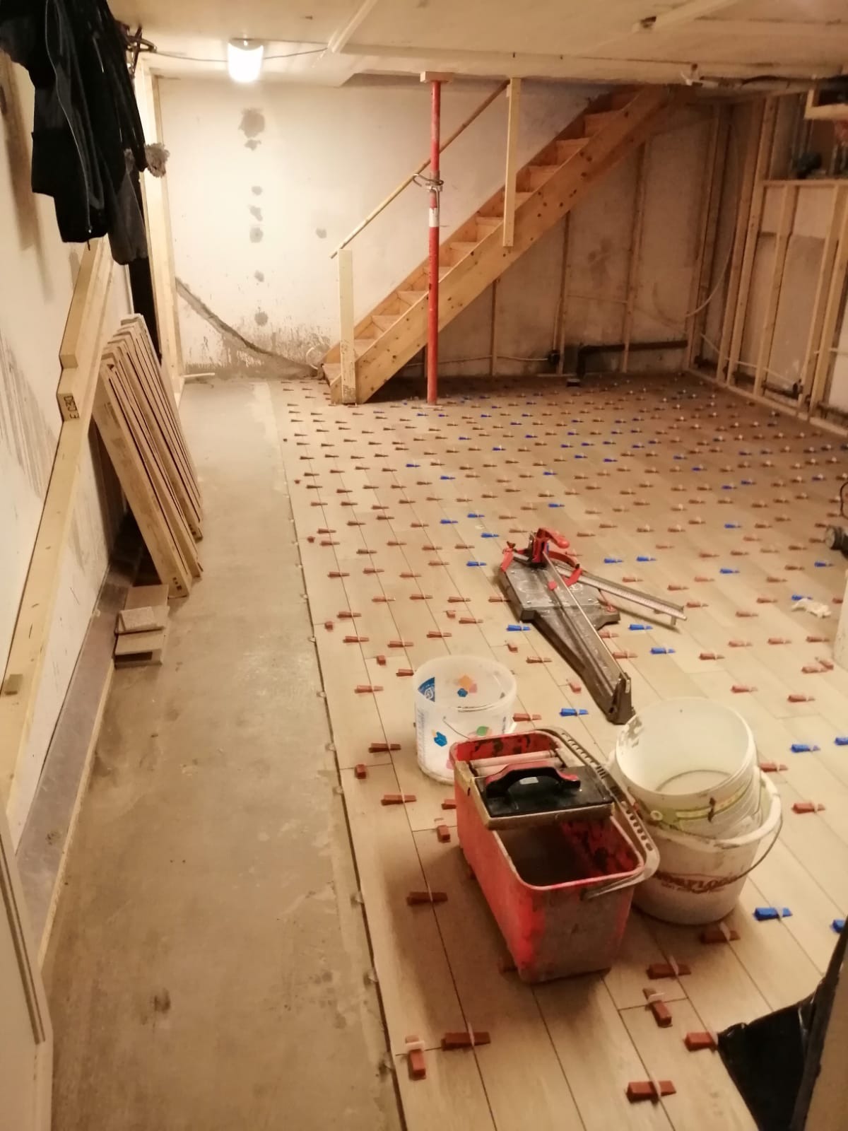 Układanie jasnych płytek podłogowych w piwnicy, z wykorzystaniem krzyżyków dystansowych, widoczna przecinarka do płytek i wiaderka po farbie.