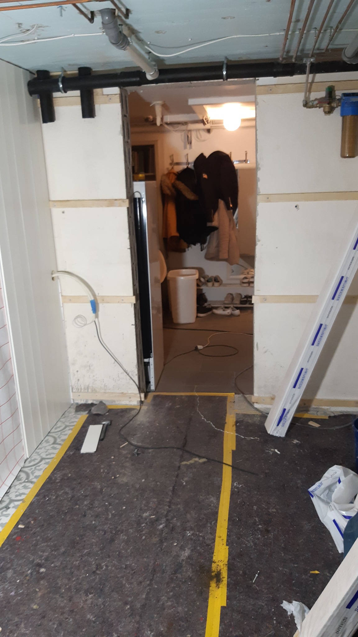 Wnętrze w trakcie remontu: widoczne przewody, rury pod sufitem, fragment ściany z drewnianymi listwami, podłoga zabezpieczona folią malarską, widok na sąsiednie pomieszczenie z garderobą i butami.